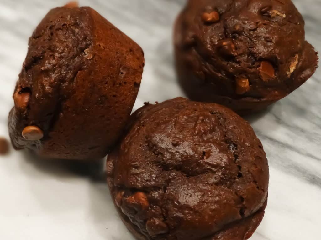 alt="three double chocolate bakery style muffins with chocolate chips on a white marble cutting board"