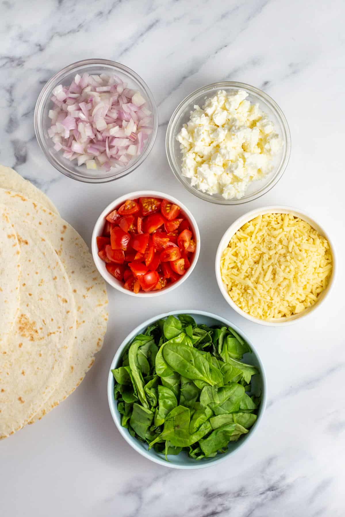 spinach, feta cheese, mozzarella cheese, red onion, tomatoes and tortilla wraps on a cutting board