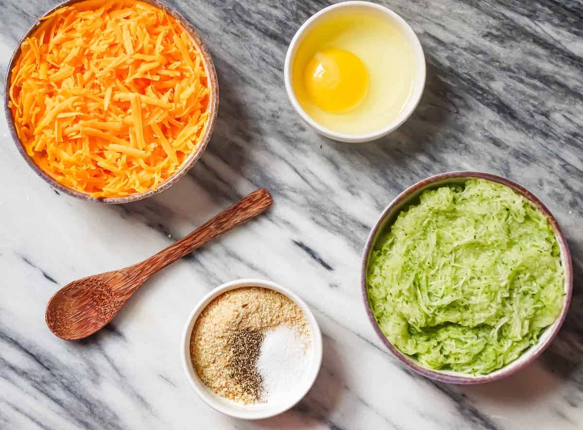 cheddar cheese, grated zucchini, salt, pepper, egg and bread crumbs on a cutting board