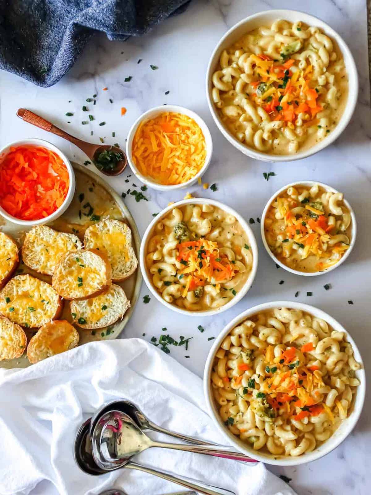 four bowls of broccoli cheddar mac and cheese with cheesy bread, carrots, and cheese for toppings