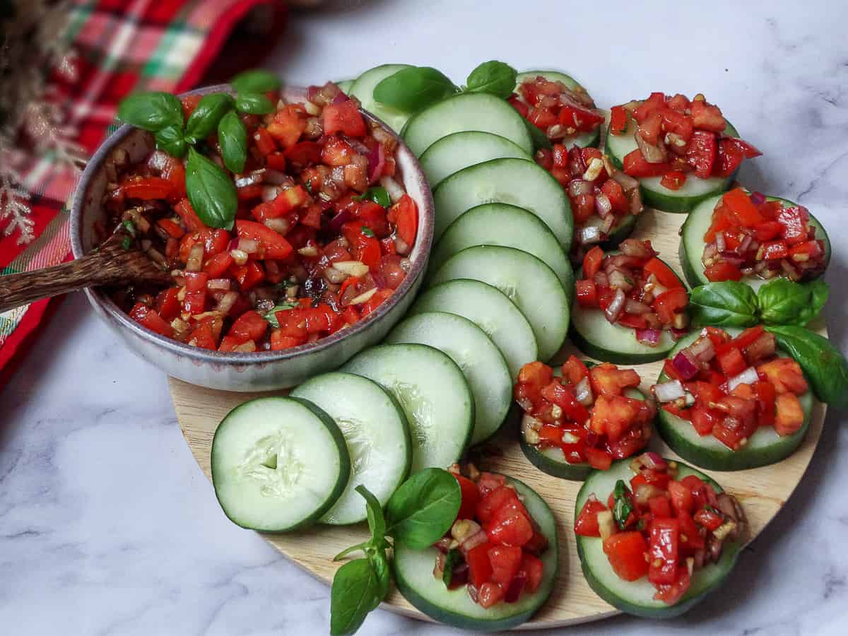 homemade bruschetta on cucumber slices arranged on a circular cutting board and topped with basil leaves served as a make ahead Christmas party appetizers and finger food