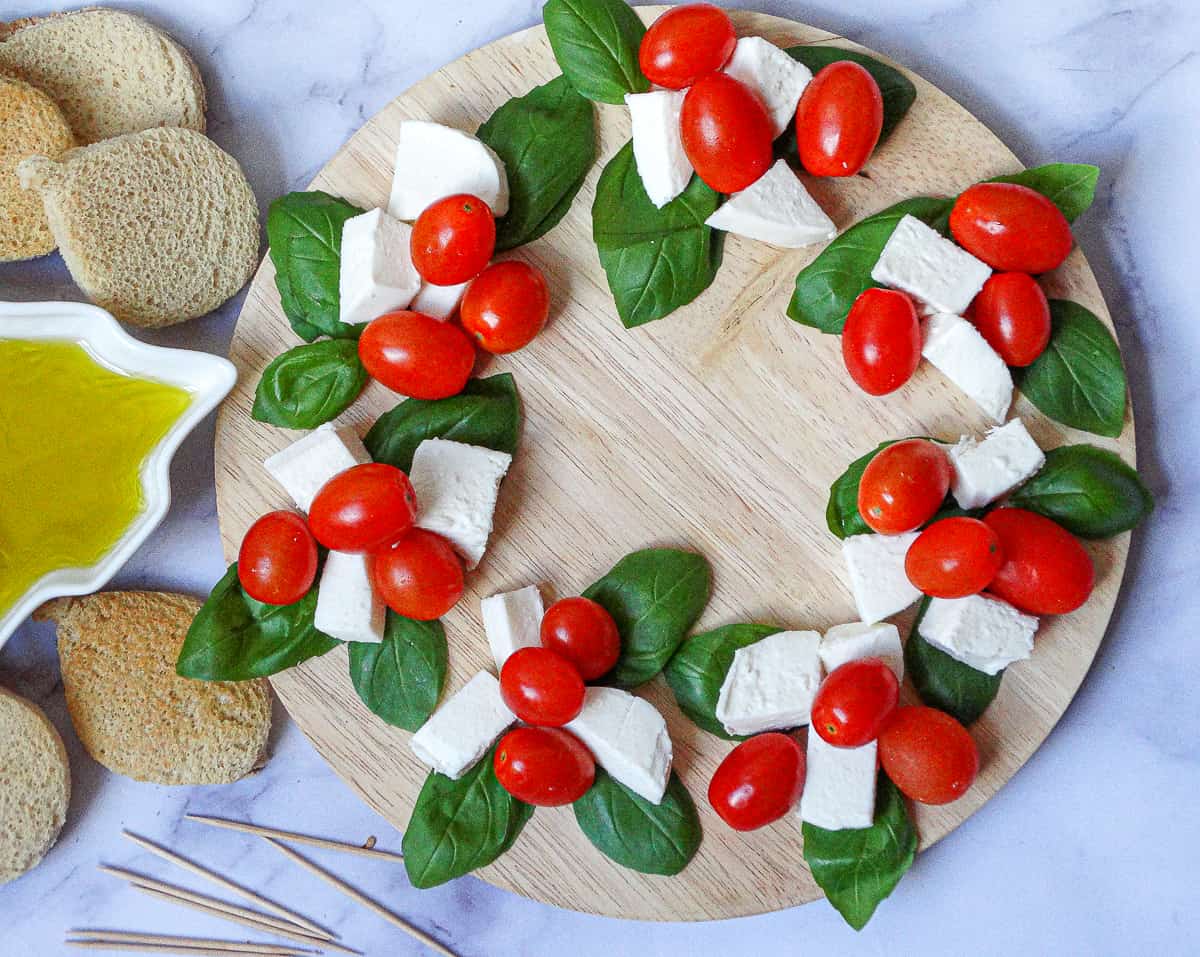 Caprese Christmas Wreath on a round wooden board, made with basil leaves, fresh mozzarella cubes and grape tomatoes
