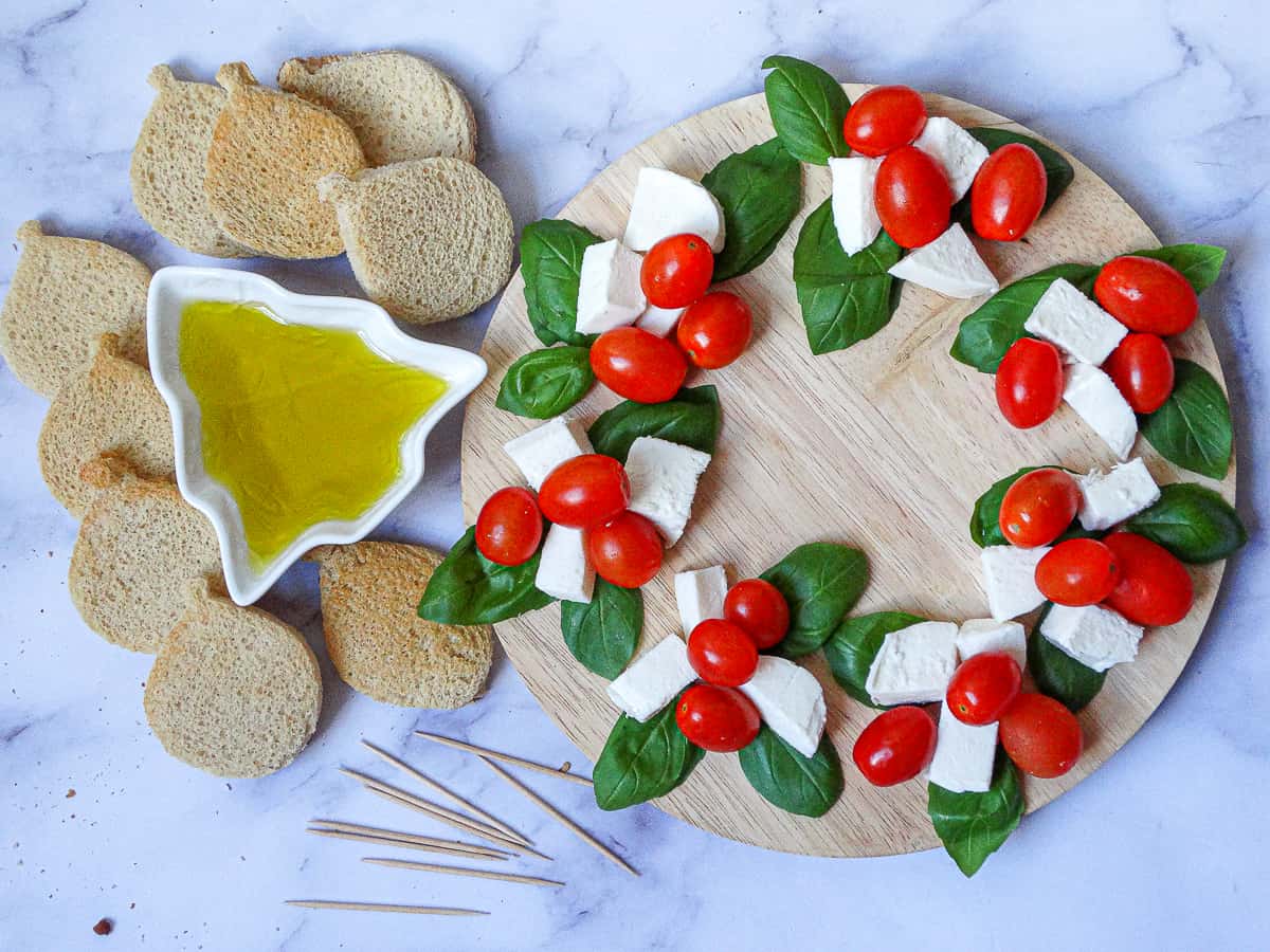 Caprese Christmas Wreath on a round wooden board, made with basil leaves, fresh mozzarella cubes and grape tomatoes