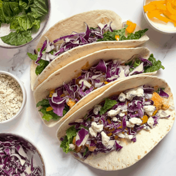 buffalo cauliflower tacos topped with red cabbage and blue cheese on a white background surrounded by fresh vegetables