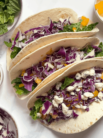 buffalo cauliflower tacos topped with red cabbage and blue cheese on a white background surrounded by fresh vegetables