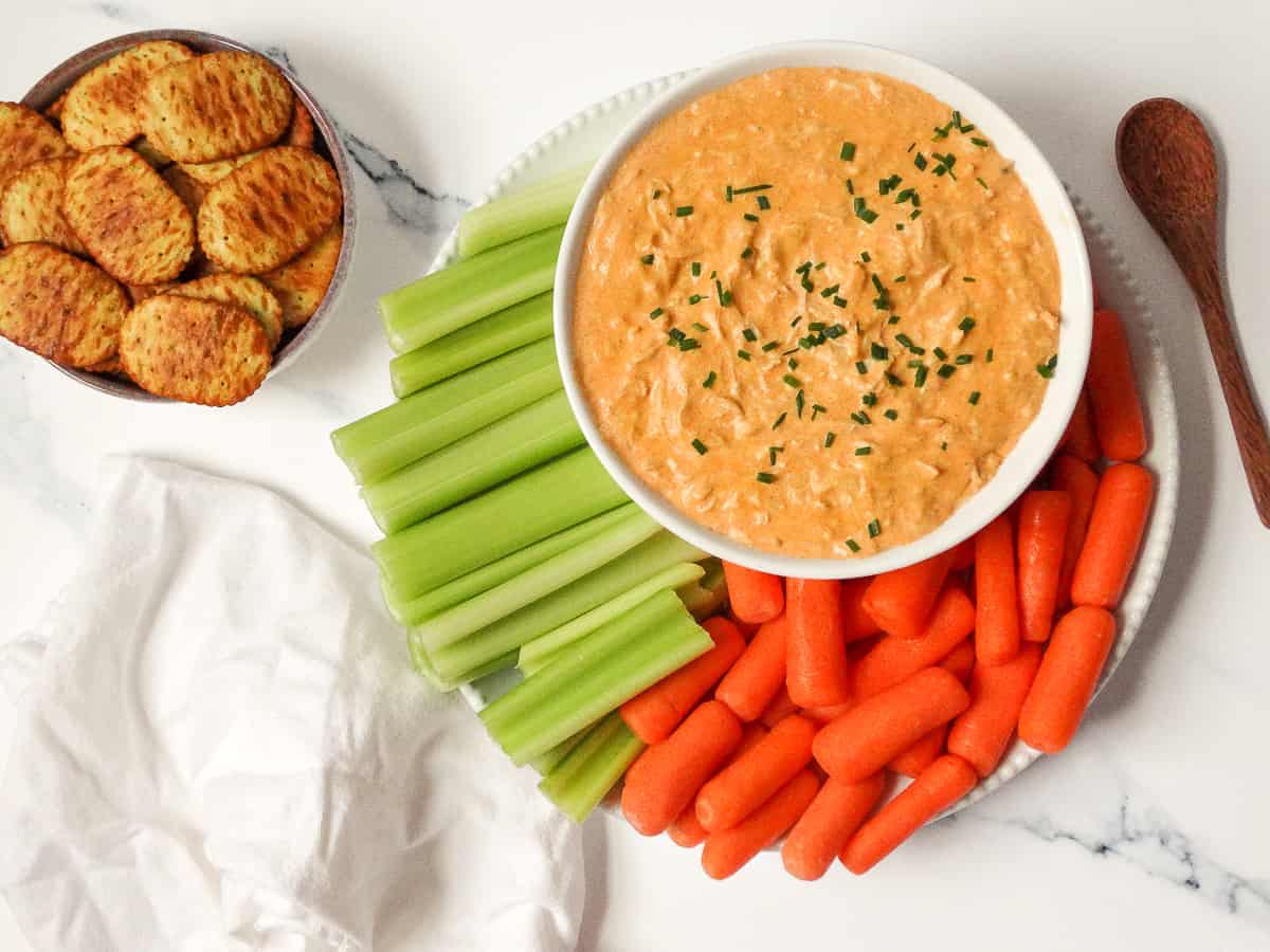 Instant Pot buffalo chicken dip topped with scallions in a white bowl surrounded by celery, carrots, and crackers