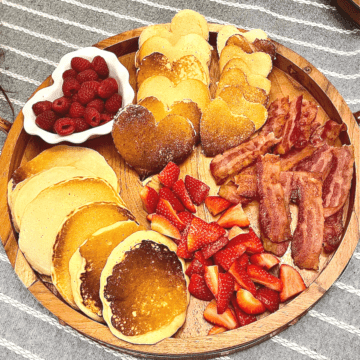 a breakfast pancake board with heart pancakes, bacon, strawberries, and raspberries