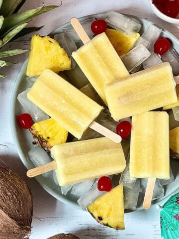 pina colada popsicles on a grey plate surrounded by cherries, pineapple slices, coconut, and decorative drink umbrellas