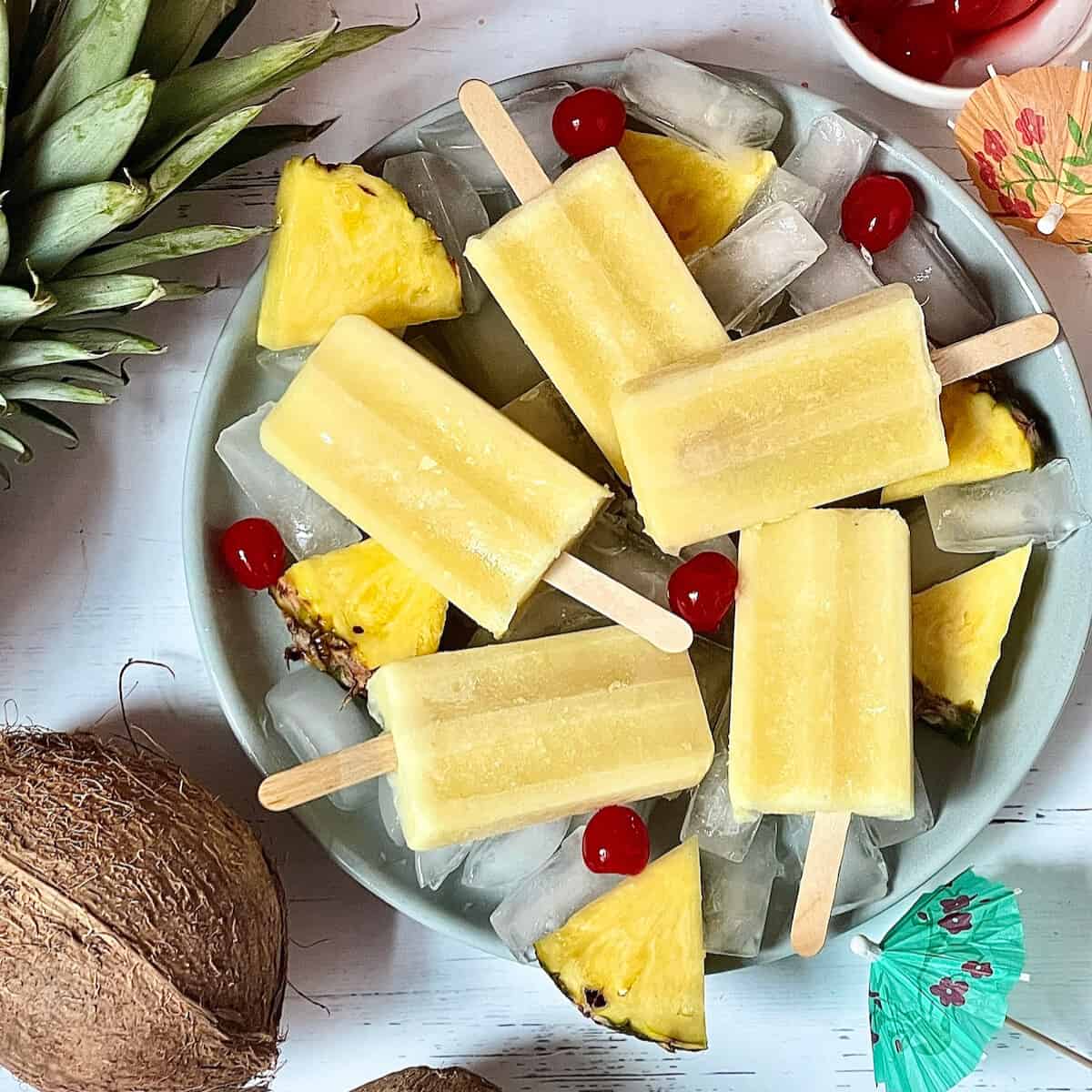 Pina colada popsicles over ice on a grey plate surrounded by cherries