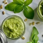 creamy pesto in 2 mason jars surrounded by pine nuts and fresh basil