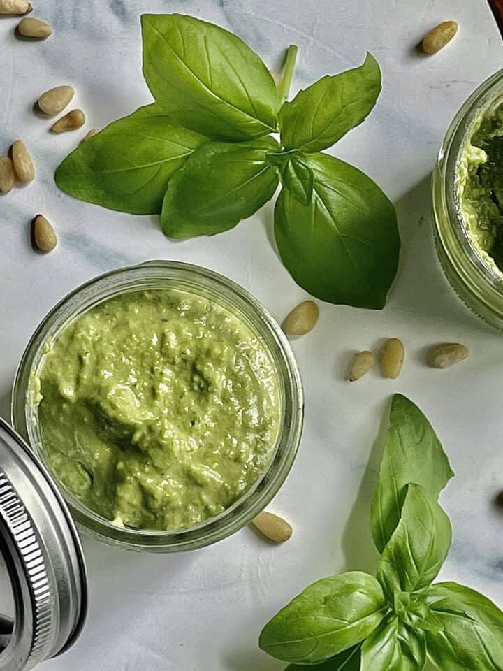 creamy pesto in 2 mason jars surrounded by pine nuts and fresh basil
