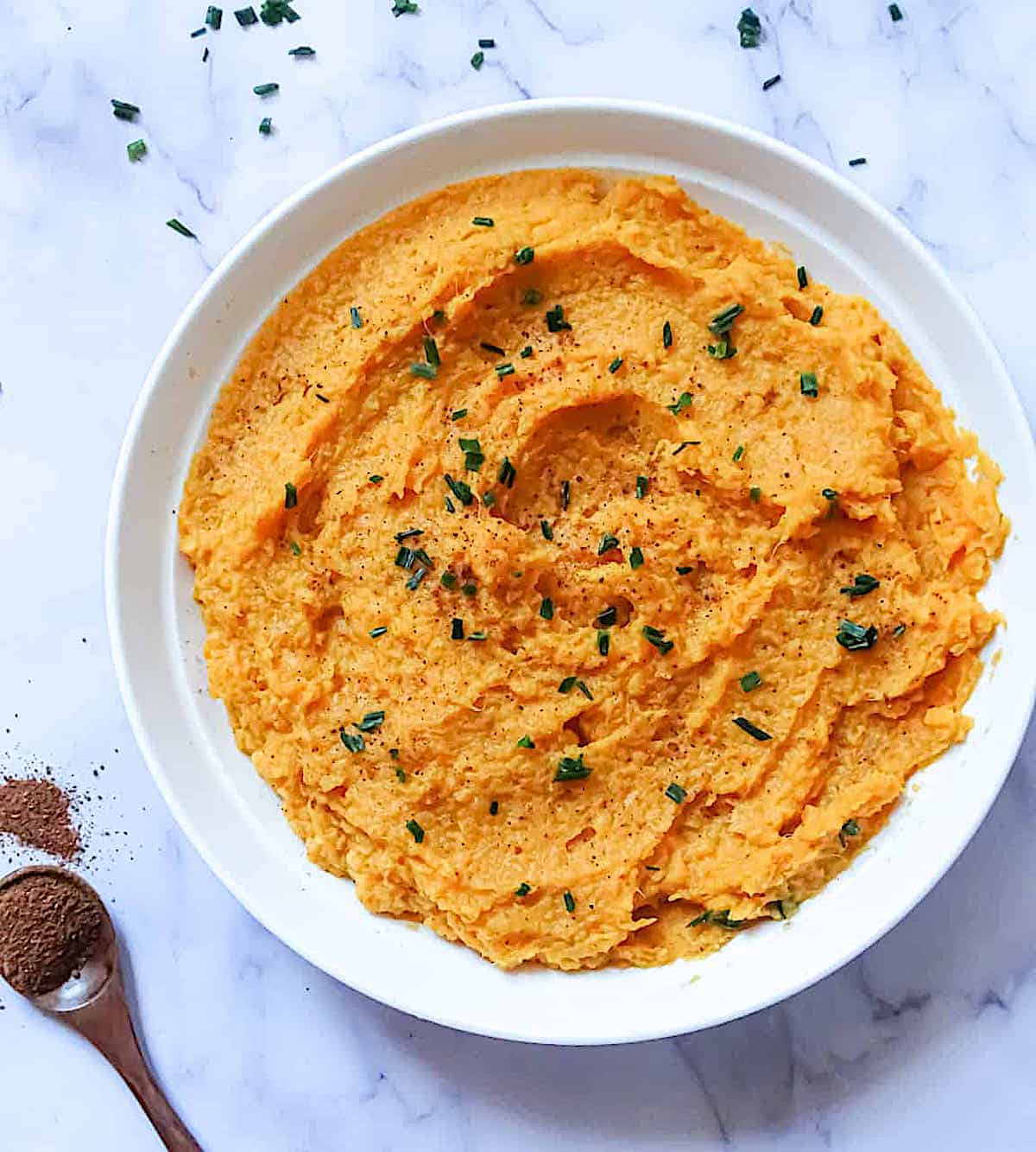 instant pot mashed sweet potatoes in a white bowl with a white background and surrounded by chives and nutmeg