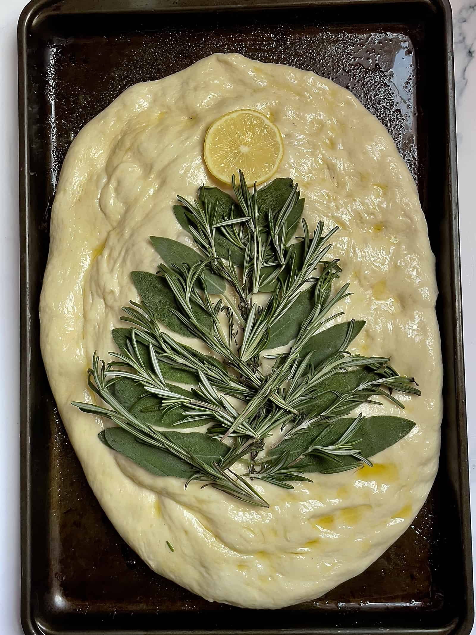 sage leaves and rosemary spread out around a piece of thyme on focaccia bread with a lemon slice as the star
