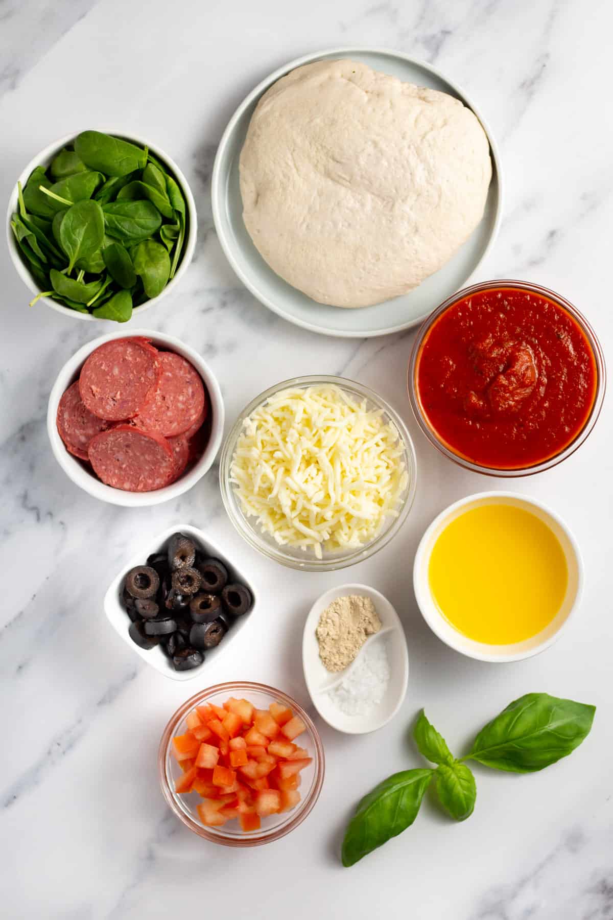 pizza dough, sauce, butter, mozzarella, pepperoni, olives, basil, salt, parsley and garlic powder in bowls on a cutting board