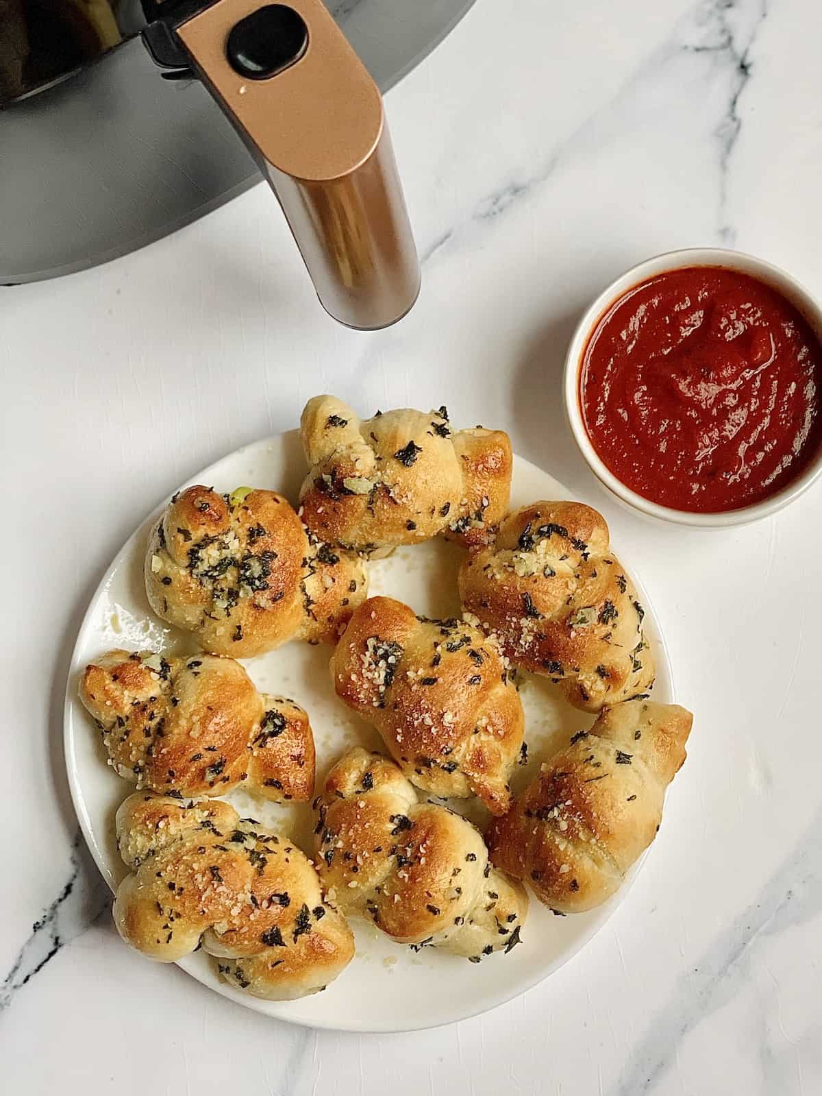 air fryer garlic knots on a white plate with dipping sauce in a small bowl next to an air fryer