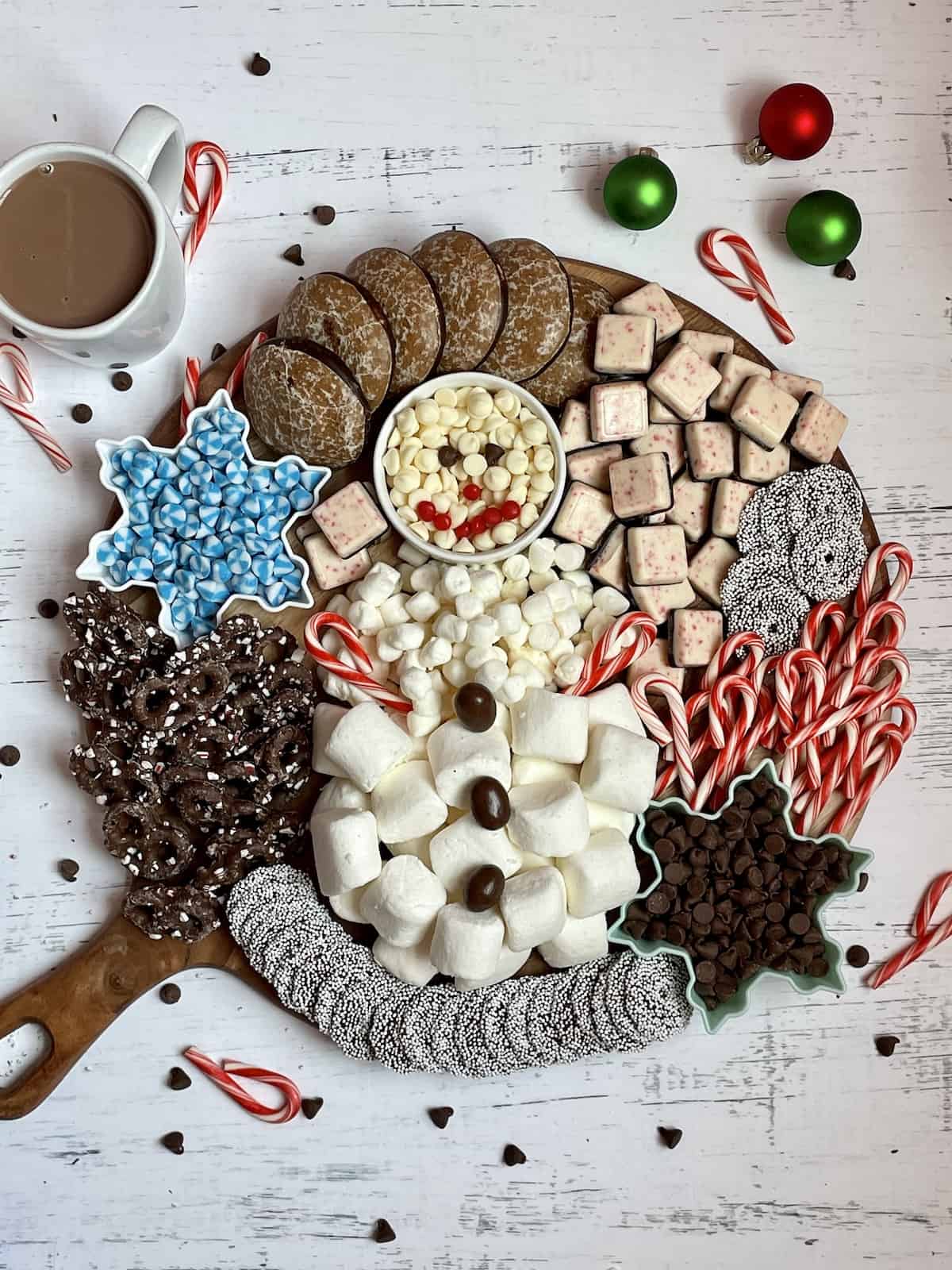 marshmallows and white chocolate chips designed in a snowman shape surrounded by candy canes, cookies, and pretzels on a wooden serving board