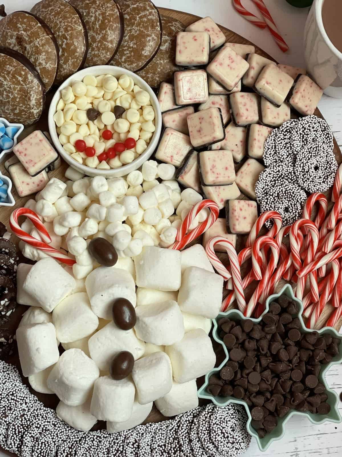 a hot chocolate charcuterie board made of marshmallows and white chocolate chips designed in a snowman shape surrounded by candy canes, cookies, and pretzels on a wooden serving board