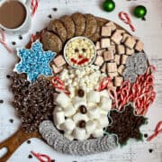 marshmallows and white chocolate chips designed in a snowman shape surrounded by candy canes, cookies, and pretzels on a wooden serving board