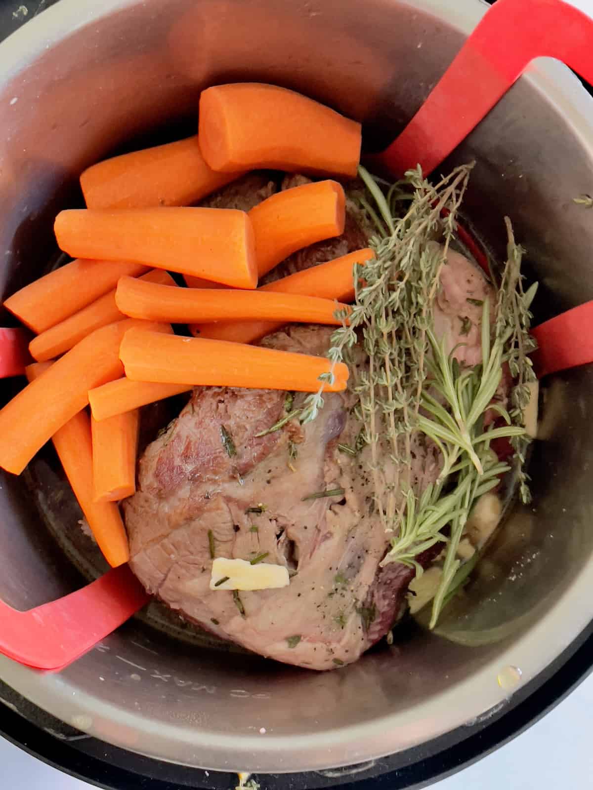 leg of lamb, carrots, broth and herb sprigs in the inner pot of a pressure cooker