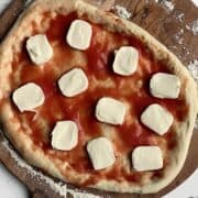 uncooked neapolitan pizza dough on a pizza peel, topped with sauce and mozzarella slices