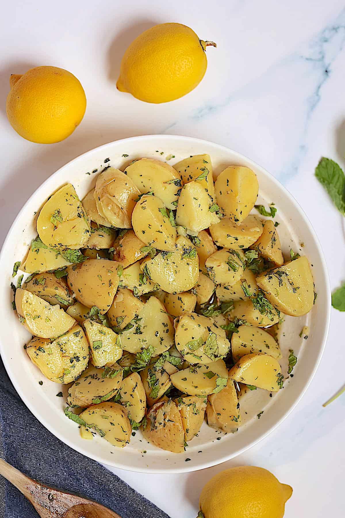 instant pot potato salad in a white bowl surrounded by lemons, mint and parsley