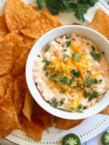 slow cooker jalapeno popper dip in a white bowl topped with cheddar cheese and cilantro, surrounded by tortilla chips on a plate